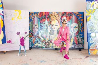 Del Kathryn Barton sits infront of a piece of her work in a pink suit with a vase of flowers to the left of her. 