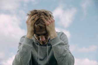 A low angle shot captures the bust and face of a young person, leaning forword and clasping his head in both hands in a sign of distress and exasperation. 