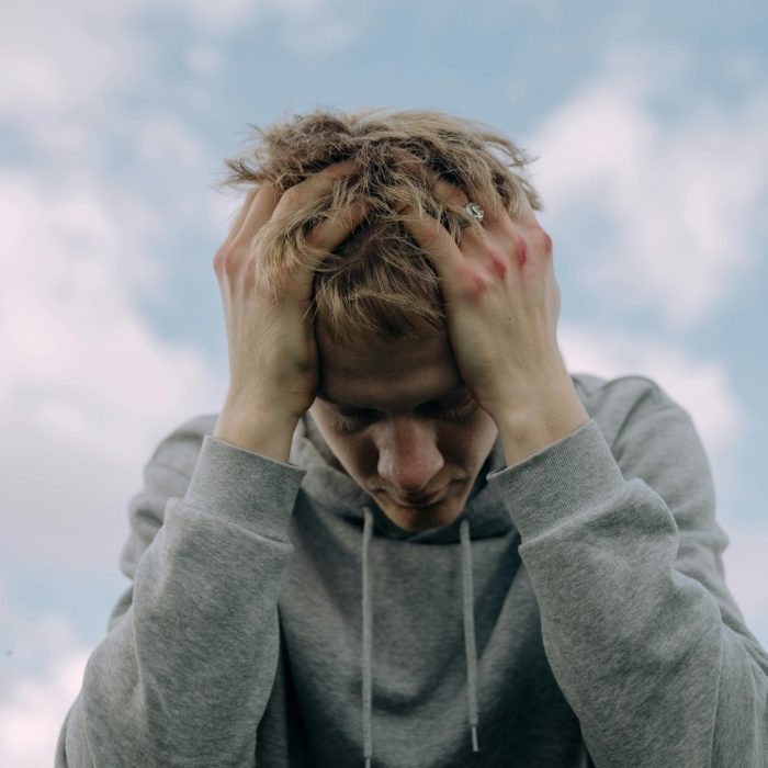 A low angle shot captures the bust and face of a young person, leaning forword and clasping his head in both hands in a sign of distress and exasperation. 