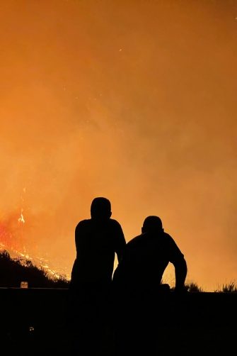 Image depicts the silhouette of two crouching figures watching a nearby bushfire engulf the landscape, burning brightly against a flaming orange sky. 