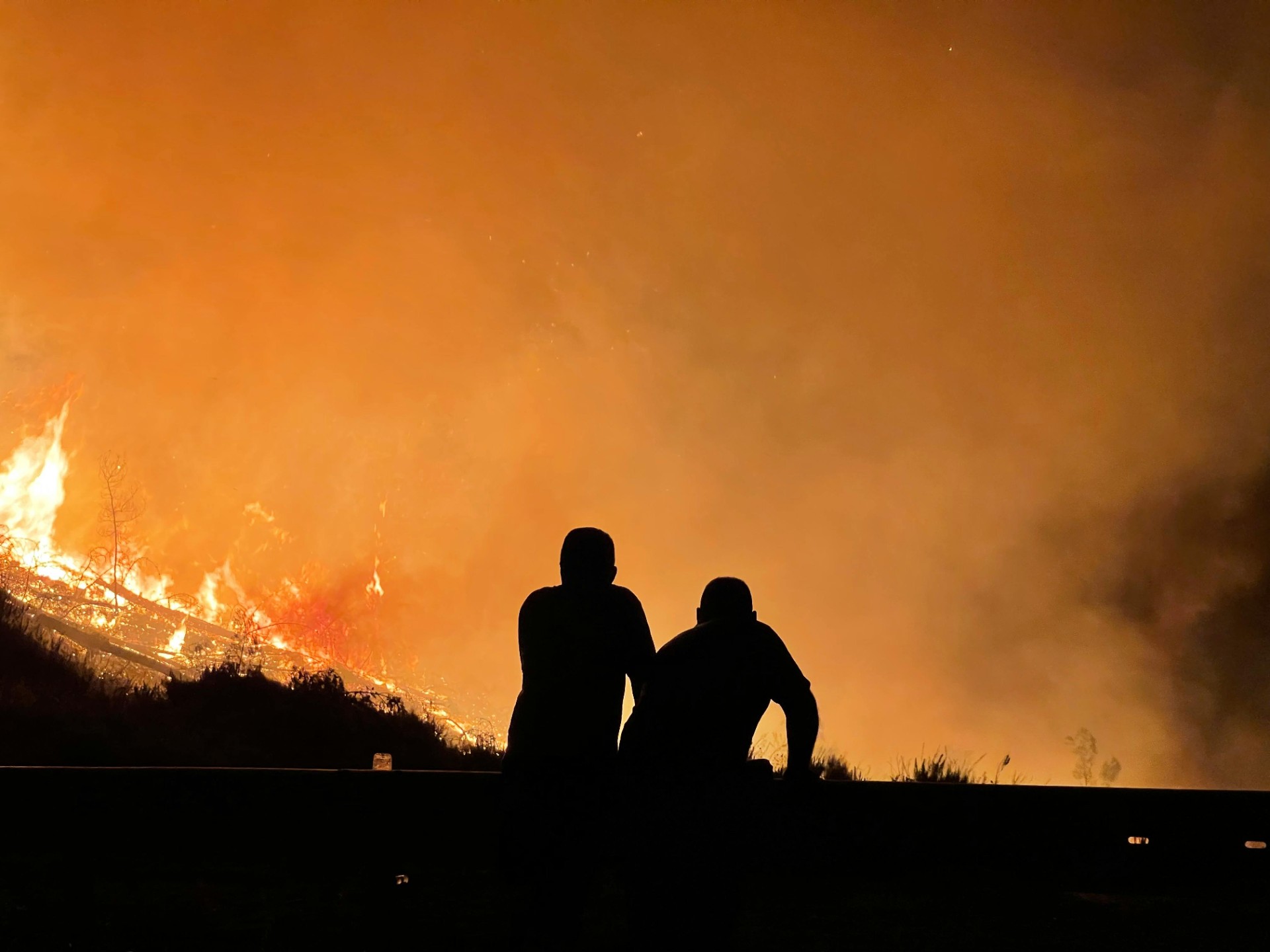 Image depicts the silhouette of two crouching figures watching a nearby bushfire engulf the landscape, burning brightly against a flaming orange sky. 