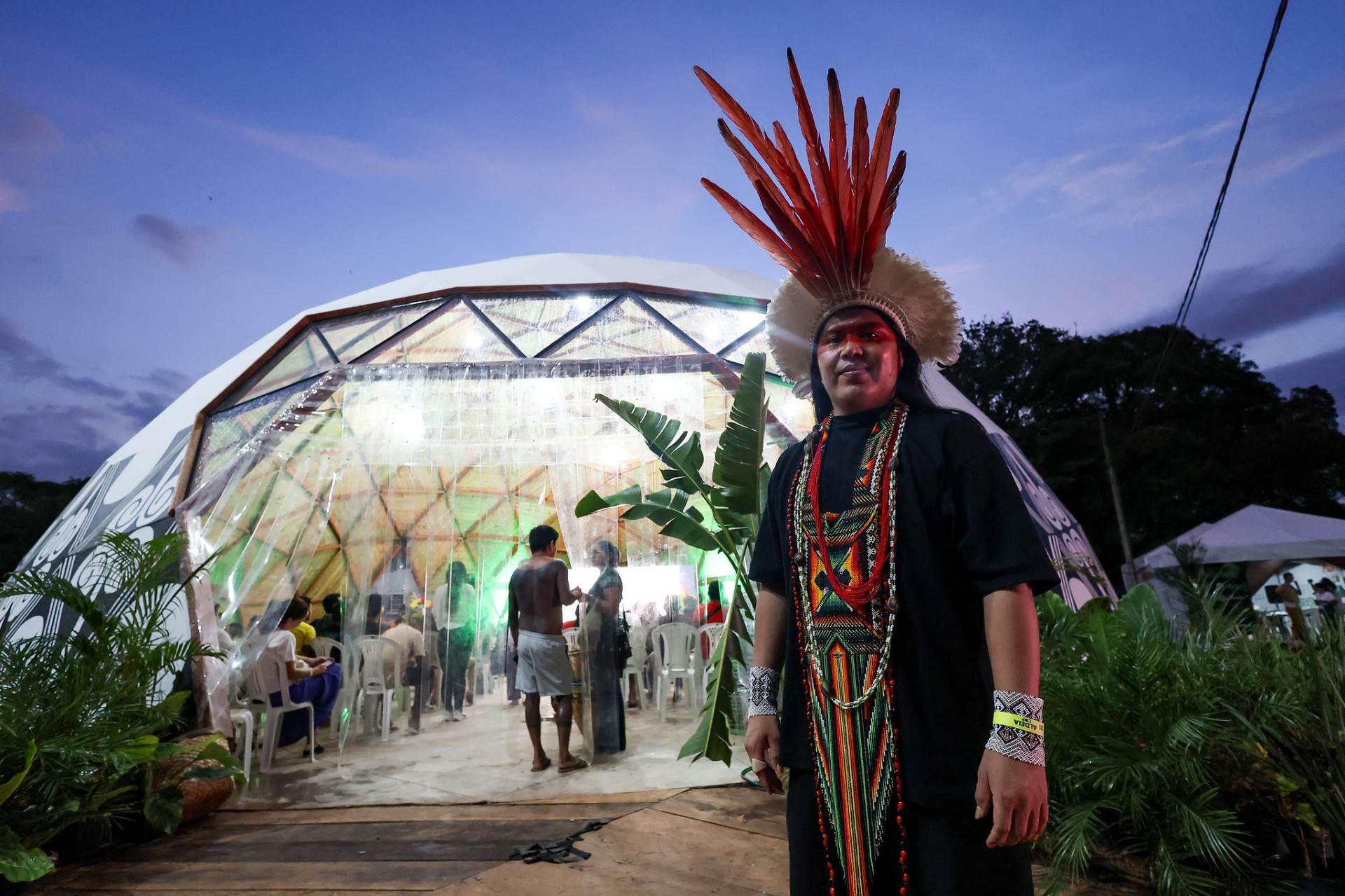 A woman in Indigenous dress stands in front of a see-through marquee shaped like a dome.