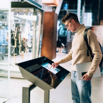 Side view of young person in glasses and with backpack standing using digital information panel in a museum