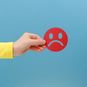Close up of hand holding round red frown face emoji paper cut out against blue background