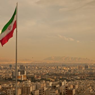Iran flag waving  in the wind above skyline of Tehran lit by orange glow of sunset.