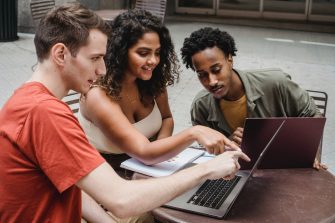 A group of three students sit together in conversation, facing an open laptop.