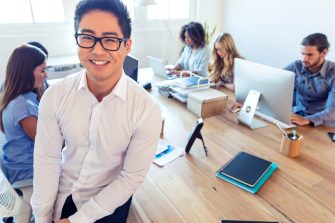Young Asian business manager standing in front of his team. Attractive business man standing in front of a table where her business team are working. There are six people in total and all are working on computers or wireless technology except the boss. All are dressed casually in a modern office setting.