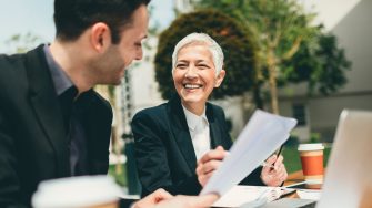 Businesspeople sitting on bench in the park and collaborating. They are having meeting out of office. Drinking coffee from disposable cup and discussing about business. Mature woman holding eyeglasses in her hand and her colleague examining documents. There is laptop on the table in front of them.