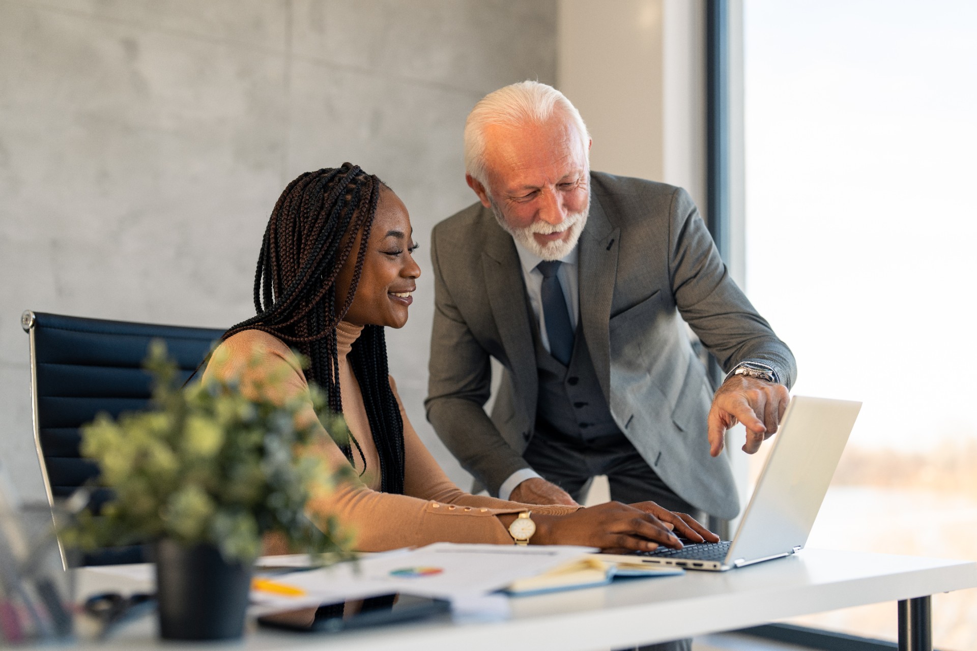 Grey haired male CEO executive manager mentor with beard giving consultation on financial operations to female African American colleague intern using laptop sitting in modern office near window.