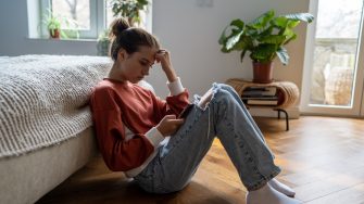 Teens and cyberbullying. Upset teen girl sitting on floor near bed using smartphone at home, scrolling social media. Child spending too much time on phone. Teenagers and gadget addiction