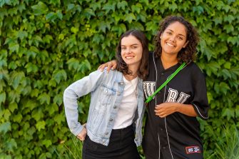 Two students with their arms around each other standing in front of a wall covered in foliage