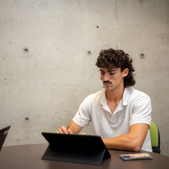 Students using their laptops at an indoor table on campus with a large blank concrete wall behind them