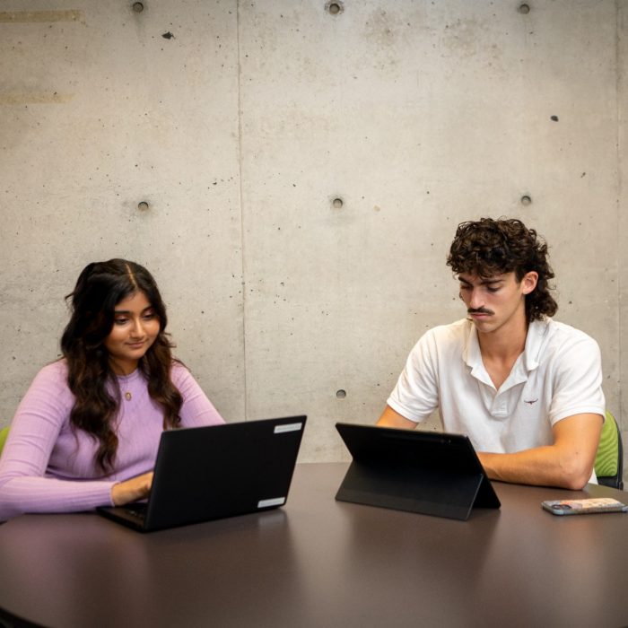 Students using their laptops at an indoor table on campus with a large blank concrete wall behind them