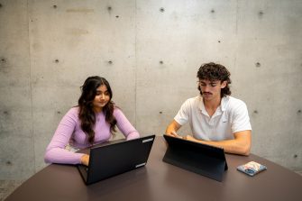 Students using their laptops at an indoor table on campus with a large blank concrete wall behind them