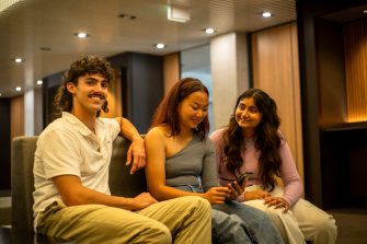 Three students smiling and looking at one of their phones while sitting on a bench indoors on campus