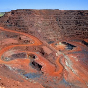 "Looking down on a large open cut iron ore mine in Australiaa's remote Pilbara region.  At the bottom of the pit at the end of the red winding road, a dump truck is being loaded with ore.  Other mine vehicles appear tiny in the huge pit.  Beyond the open cut are green hills.  Taken with permission during a guided tour of the mine."