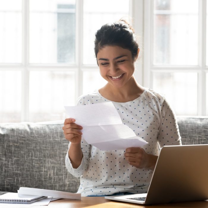 Woman sitting on couch at home reading paper notice receive good news feels happy, cheerful student female looking at document enjoy exam results or college admission letter concept