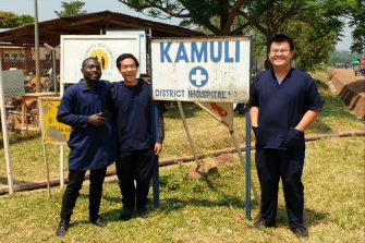 Three people standing with the Kamuli District hospital sign