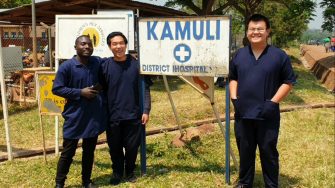 Three people standing with the Kamuli District hospital sign
