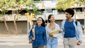 Three diverse college friends walking arm in arm, laughing and carrying books and backpacks on a sunny day.