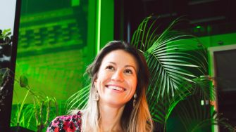 A woman sits with a laptop on her knee, smiling off camera