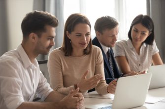 Female manager mentor teach help male intern trainee new employee explain online strategy on laptop at group meeting, focused business woman talk to colleague client work together cooperate in office