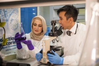 male and female student at micro biomedical research centre looking at microscope and glass jar wearing protective lab equipment