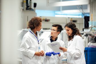 Diverse mix of technicians, wearing lab coats, debate working procedure, in a disease research facility. Nuclear magnates resonance equipment, is in the background, this high end apparatus can be used to discover information on conditions such as dementia, diabetes, autism and cancer. Focus technique and layers, ensure the group of female science graduates and manager are the centre of attention. This is a realistic scenario.