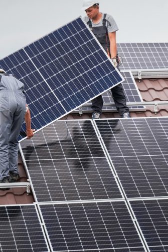 Technicians in blue suits mounting photovoltaic solar panels on roof of modern house. Solar modules as ecological renewable energy sources. Alternative production modules power sustainable resources