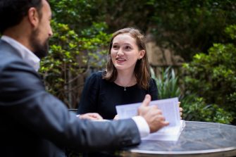 A woman looks inspired while discussing the contents of a book that a man has out on the table where they are seated at an outdoor cafe.