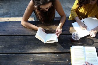 Three women are seated at a lunch table reading from notebooks.