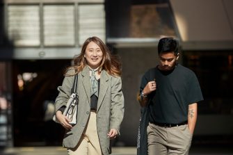 Female in green jacket and male in black t-shirt, law students, walking together behind the law building.  Shadows and light on buildinf exterior.