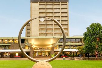 Looking through the large circular clock statue on the lawn towards the multi-storey Main Library building in the distance.. Golden light shows under the large white entrance ceiling and a clear light blue sky behind the building.