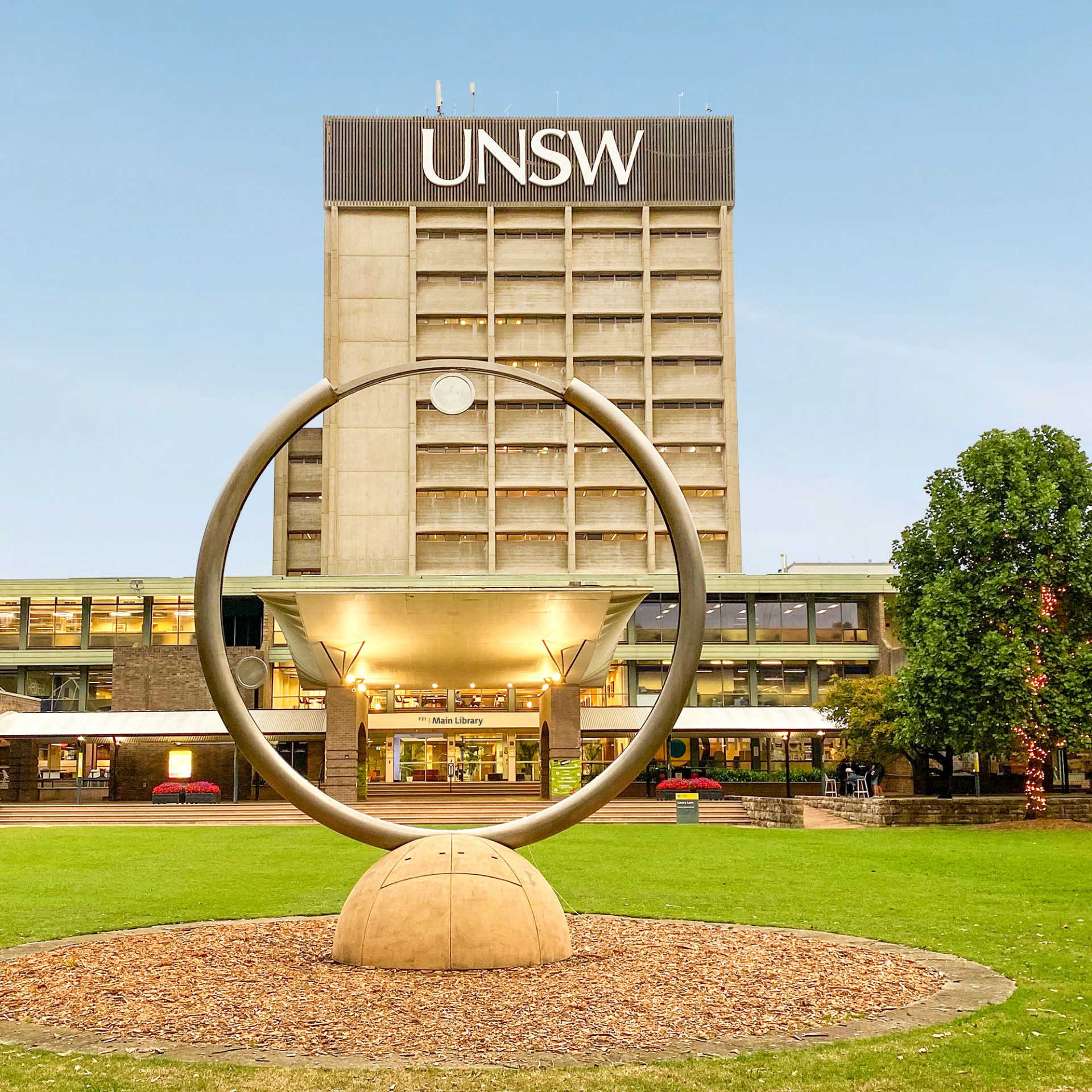 Looking through the large circular clock statue on the lawn towards the multi-storey Main Library building in the distance.. Golden light shows under the large white entrance ceiling and a clear light blue sky behind the building.