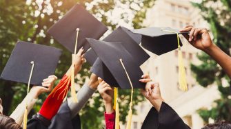 6 arms high in the air, most with black graduation gown sleeves showing, hold black graduation mortar board hats. Trees and building in the distant background.