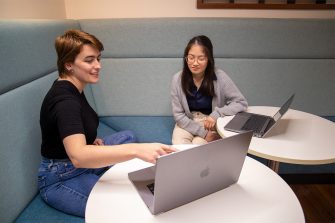 Two women seated inside the Library. One points at a laptop while the other looks towards same laptop screen.