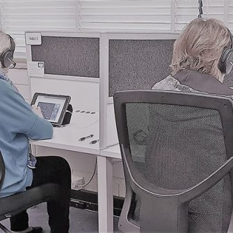 Two individuals seated at separate workstations, each using a tablet and wearing headphones. The stations are divided by a partition, with a window and white brick wall in the background