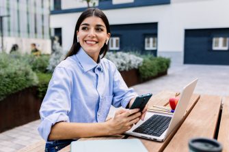 Happy entrepreneur woman with laptop using cell phone sitting outside of office building. Business lifestyle concept.