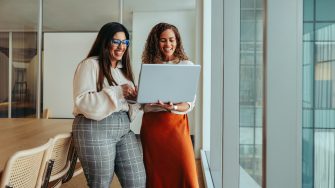 Two businesswomen working as a team in a tech startup environment. They are reviewing information on a laptop, reflecting a collaborative and innovative workplace atmosphere.
