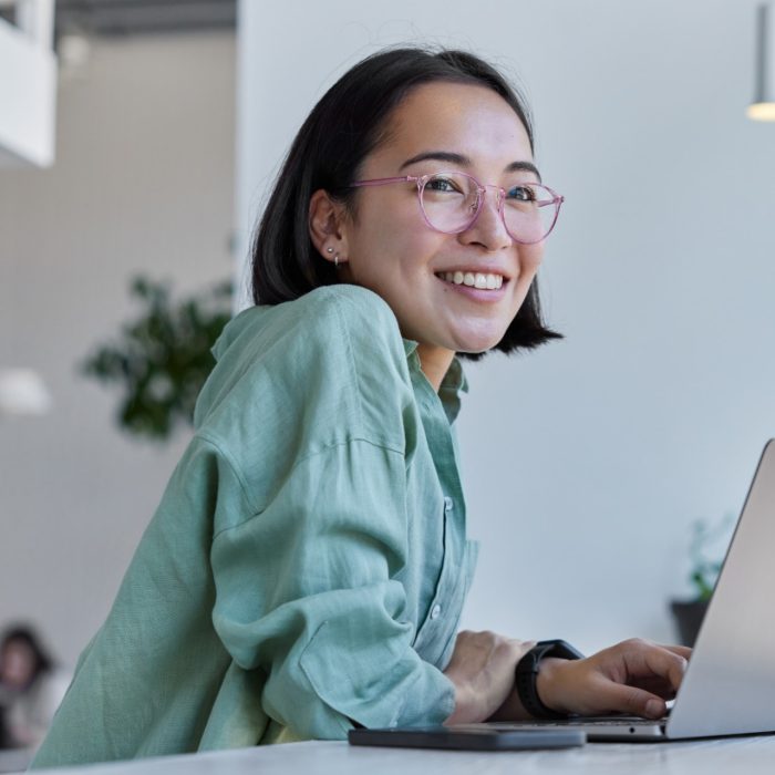 Pretty cheerful Asian woman in eyeglasses and casual clothes browses laptop computer connected to 4g internet updates software uses modern technologies poses in cafeteria looks gladfully away