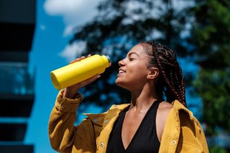 Attractive young woman drinking from a yellow bottle. She is wearing a yellow jacket and it is a hot day in summer. Smiling. Mixed race.