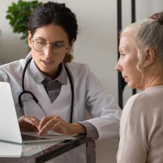 Young female doctor use computer consult mature patient in modern hospital. Woman GP and senior client look at laptop screen, discuss treatment or therapy in clinic. Elderly healthcare concept.