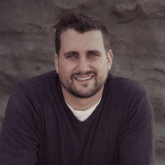 Portrait of Paul Munro wearing a dark jumper with white t-shirt. Paul has short hair and a neatly trimmed beard.