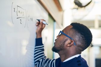 Man with glasses in striped jumper writing mathematical equation on whiteboard