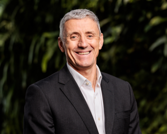 A headshot of Dr Nicholas Wood smiling in a black suit and white shirt standing in front of a blurred out green wall