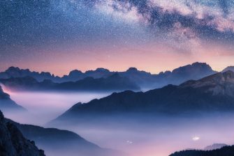 Milky Way over mountains in fog at night in summer. Landscape with foggy alpine mountain valley, purple low clouds, colorful starry sky with milky way, city illumination. Dolomites, Italy. Space