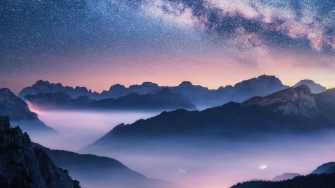 Milky Way over mountains in fog at night in summer. Landscape with foggy alpine mountain valley, purple low clouds, colorful starry sky with milky way, city illumination. Dolomites, Italy. Space