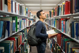Student with backpack and open book standing between book shelves in library