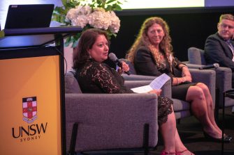 A woman is sitting on a stage beside other guests speaking into a microphone.