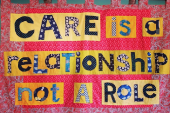 A handmade pink material banner held up by a curtain pole with the words 'Care is a Relationship not a Role' patchworked on to a yellow background